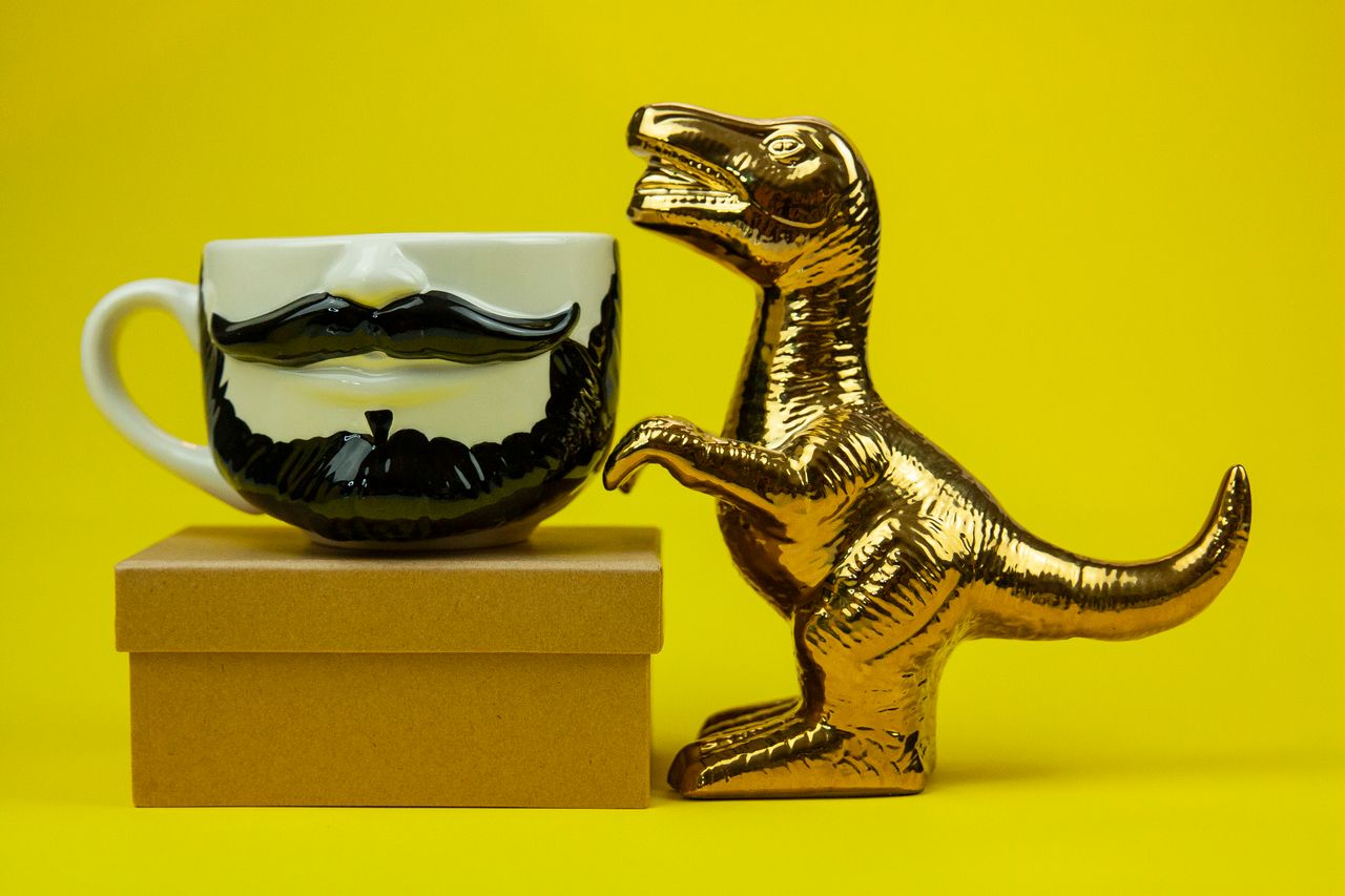 A whimsical gold dinosaur figurine and mustache mug displayed on a box against a bright yellow background.